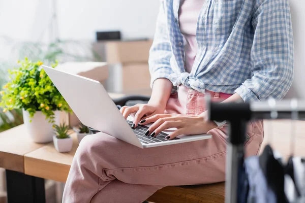 Cropped view of volunteer using laptop while sitting near hanging rack and plants in charity center — Stock Photo