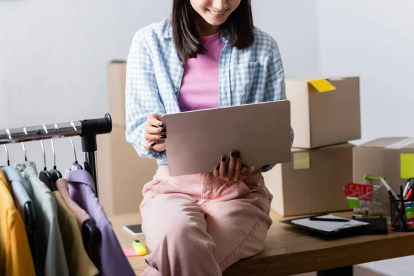 Vista ritagliata del manager sorridente che tiene il computer portatile vicino a scatole e vestiti su rack appeso in primo piano sfocato nel centro di beneficenza — Foto stock