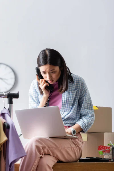 Asiatico manager parlando al telefono e utilizzando laptop vicino scatole di cartone e vestiti su appendiabiti — Foto stock
