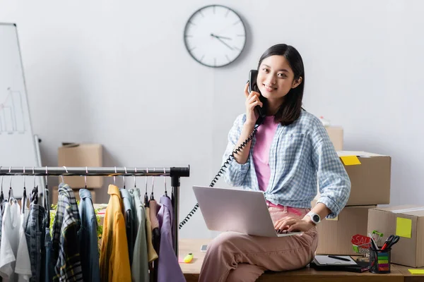 Smiling asian volunteer talking on telephone and using laptop near clothes and cardboard boxes in charity center — Stock Photo