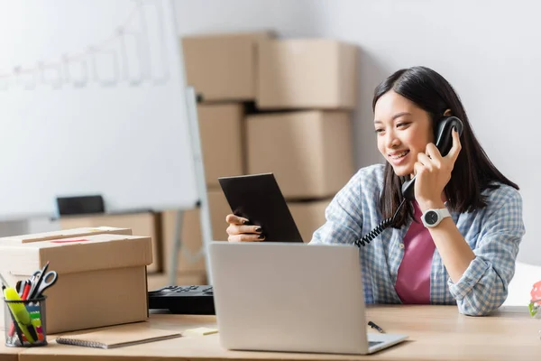 Smiling asian volunteer talking on telephone and looking at clipboard near laptop and carton box on blurred foreground — Stock Photo