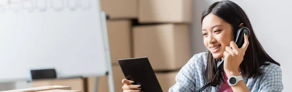 Smiling asian volunteer looking at clipboard and talking on telephone in charity center, banner — Stock Photo