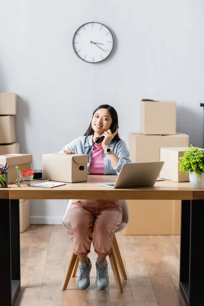 Positive asian manager talking on telephone near laptop and carton boxes on table in charity center — Stock Photo