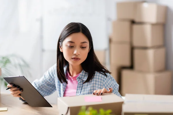 Asian volunteer holding clipboard while looking at cardboard box on blurred foreground — Stock Photo