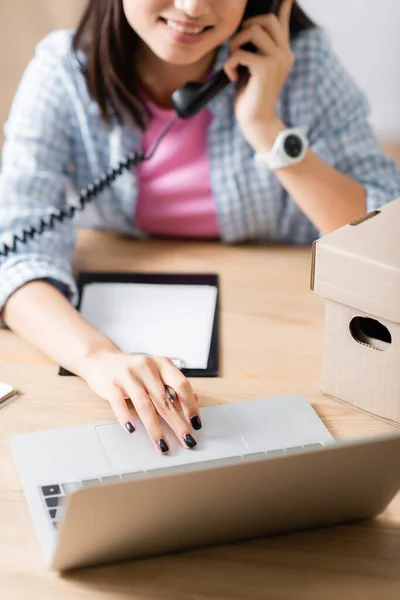 Vista cortada do voluntário sorrindo usando laptop enquanto conversa no telefone perto do pacote no fundo embaçado — Fotografia de Stock