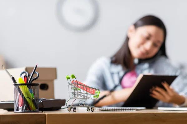 Carrito de compras de juguete con letras de venta en etiqueta de precio cerca de papelería y voluntario hablando por teléfono sobre fondo borroso - foto de stock