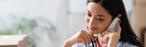 Lächelnder asiatischer Manager hält Stift in der Hand, während er im Charity Center mit dem Smartphone spricht, Banner — Stockfoto