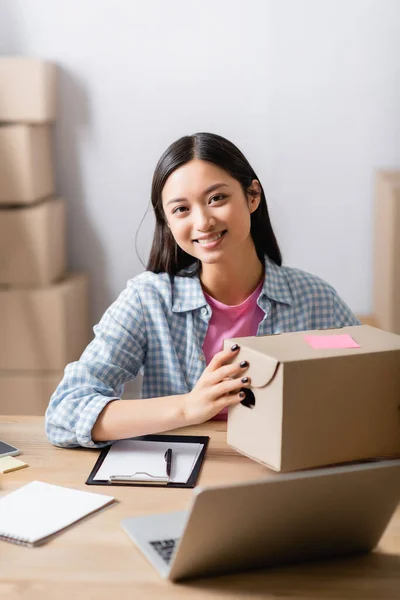 Voluntario asiático mirando a la cámara mientras sostiene la caja cerca del portapapeles y el portátil en primer plano borroso - foto de stock