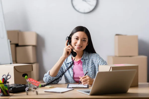 Smiling asian volunteer talking on telephone near clipboard, toy shopping cart and laptop on blurred foreground in charity center — Stock Photo