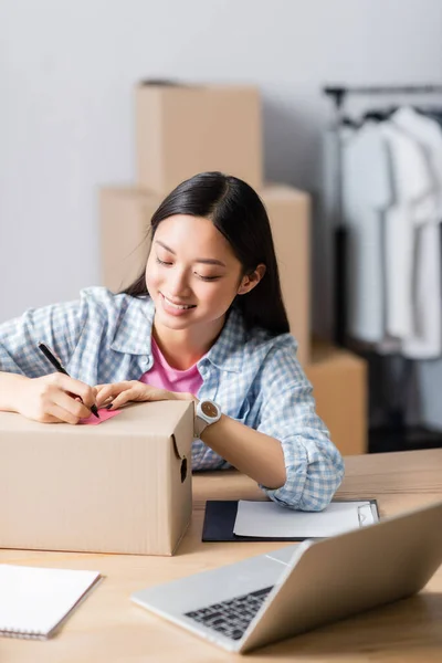 Smiling asian volunteer writing on sticky note on carton box near clipboard and laptop on blurred foreground — Stock Photo