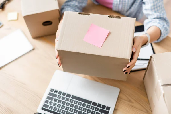 Cropped view of volunteer holding box with sticky note near laptop on blurred background — Stock Photo