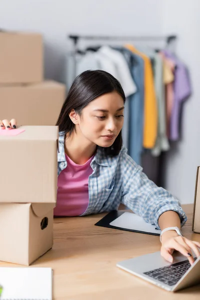 Asian volunteer using laptop near boxes on blurred foreground in charity center — Stock Photo