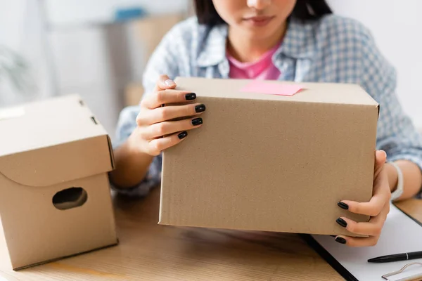 Cropped view of volunteer holding carton box with sticky note near clipboard and pen in charity center — Stock Photo