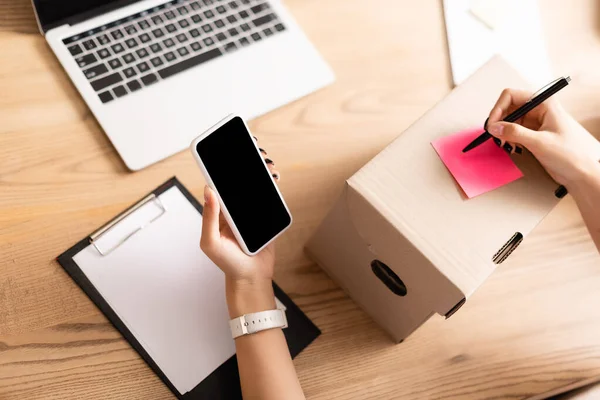 Cropped view of woman holding smartphone with blank screen while writing on sticky note on box near laptop in charity center — Stock Photo