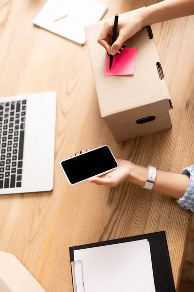 Cropped view of volunteer holding smartphone with bank screen while writing on box with sticky note near laptop on blurred background — Stock Photo