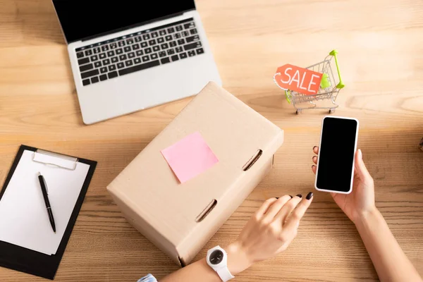 Top view of volunteer holding smartphone with blank screen near box, clipboard and toy shopping cart with sale lettering — Stock Photo