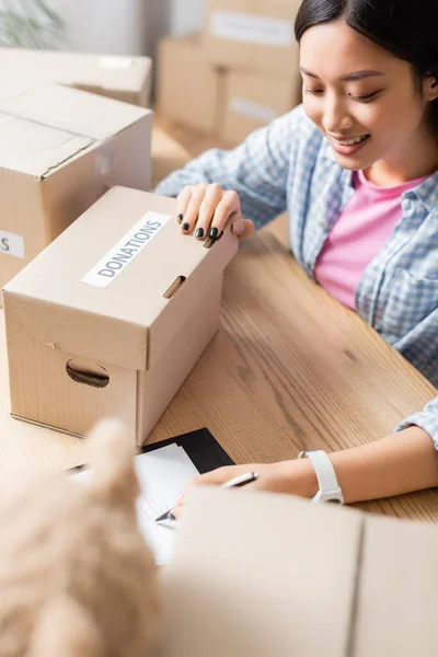 Smiling asian volunteer writing on clipboard near boxes with donations lettering and soft toy on blurred foreground in charity center — Stock Photo