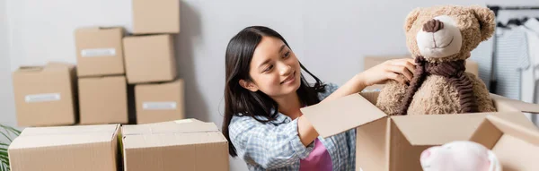 Positive asiatische Freiwillige legen Plüschtier in Schachtel in der Nähe von Paketen auf unscharfen Vordergrund, Banner — Stockfoto