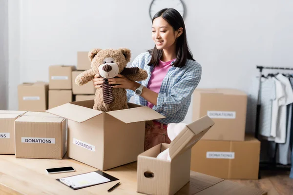 Asian volunteer holding soft toy near boxes with donations lettering, clipboard and smartphone in charity center — Stock Photo