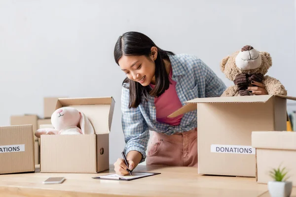 Smiling asian volunteer writing on clipboard near boxes with donations lettering and soft toys in charity center — Stock Photo