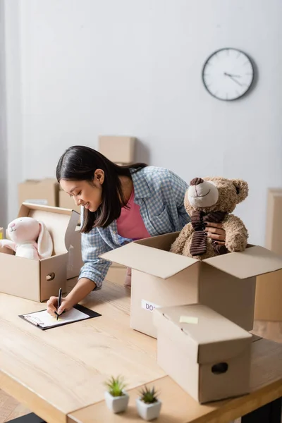 Smiling asian volunteer writing on clipboard near packages with soft toys in charity center — Stock Photo