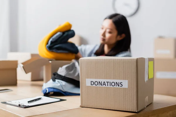 Box with donations lettering near asian woman holding clothes in charity center on blurred background — Stock Photo
