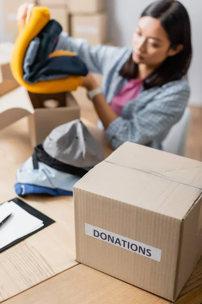 Package with donations lettering near clipboard and asian volunteer with clothes on blurred background — Stock Photo