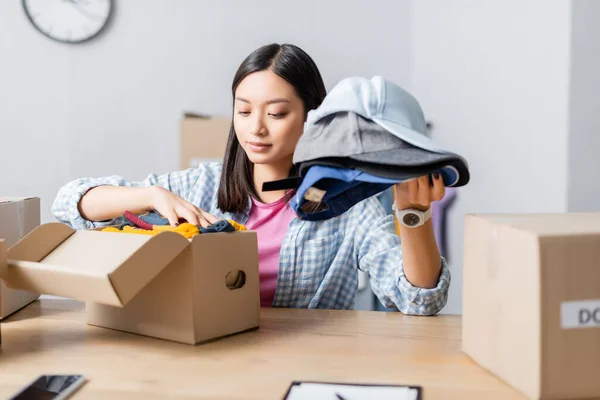 Asiatico donna putting clothes in carton box near smartphone su blurred foreground in charity center — Foto stock