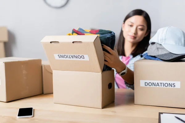 Smartphone near boxes with donations lettering and asian volunteer holding clothes on blurred background — Stock Photo