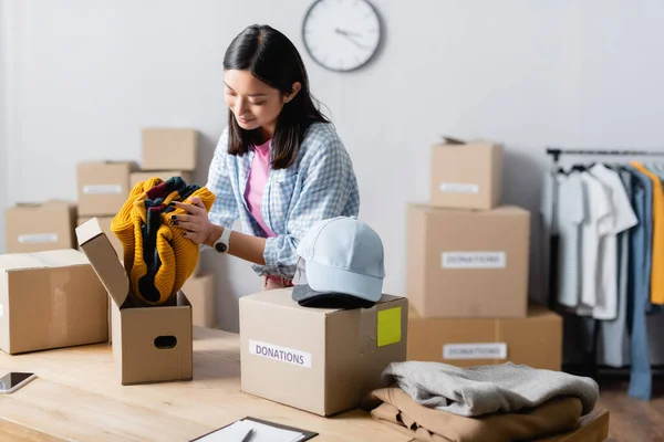Asian manager of charity center putting clothes near boxes with donations lettering and smartphone while working in charity center — Stock Photo