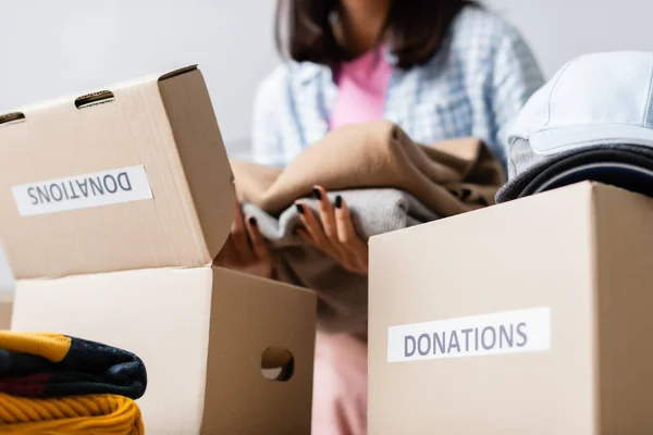 Carton packages with donations lettering near young volunteer with clothes on blurred background — Stock Photo