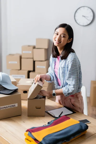 Smiling asian volunteer packing clothes in box near smartphone and clipboard in charity center — Stock Photo