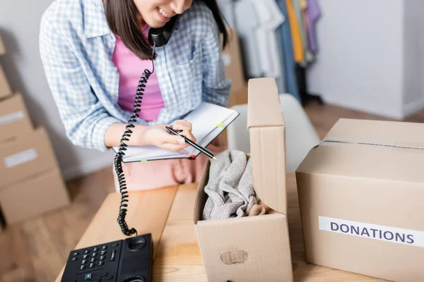 Cropped view of woman talking on telephone and pointing at clothes in box with donations in charity center — Stock Photo