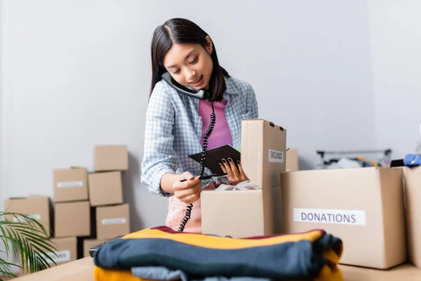 Asian woman holding notebook and talking on telephone near boxes with donations and clothes on blurred foreground in charity office — Stock Photo