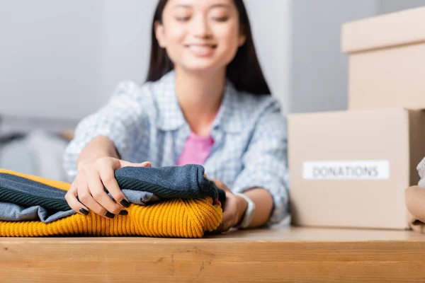 Sonriente asiático voluntario sosteniendo ropa cerca de cajas de cartón en borrosa fondo - foto de stock