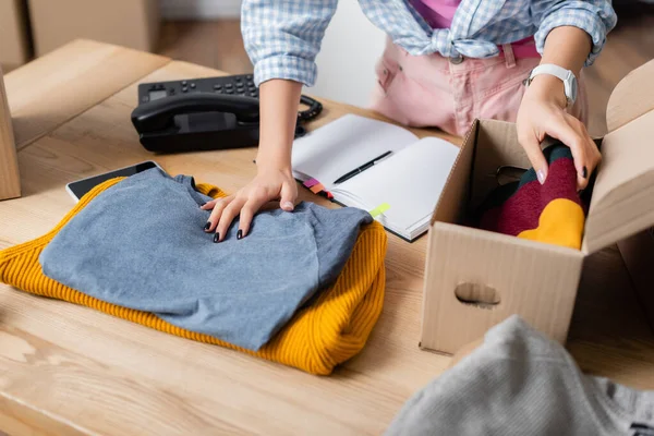 Cropped view of volunteer holding clothes near box, smartphone and notebook on table — Stock Photo