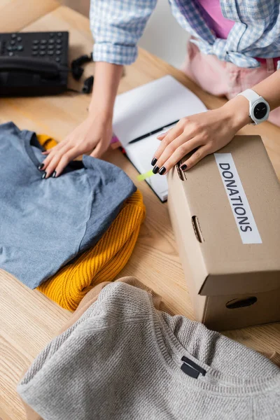 Cropped view of volunteer standing near clothes, box with donations lettering and notebook on blurred background — Stock Photo