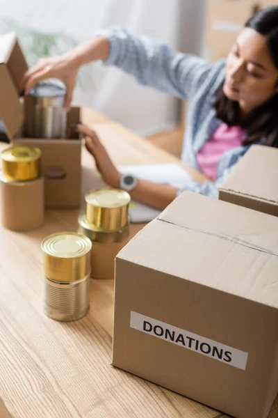 Box with donations lettering near canned food and asian volunteer on blurred background — Stock Photo