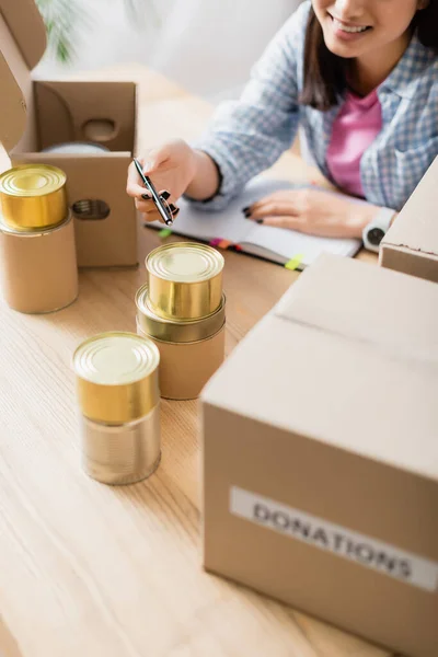 Vista recortada de un voluntario sonriente sosteniendo la pluma cerca de alimentos enlatados y cajas en primer plano borroso - foto de stock