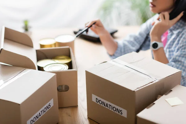 Cropped view of boxes with donations lettering and tin cans on table near volunteer talking on telephone on blurred background — Stock Photo