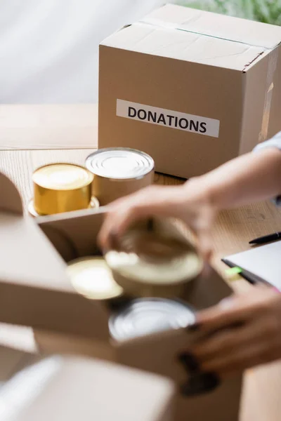 Ausgeschnittene Ansicht der Verpackung mit Spendenaufdruck in der Nähe von Blechdosen und freiwilligen Helfern im unscharfen Vordergrund — Stockfoto