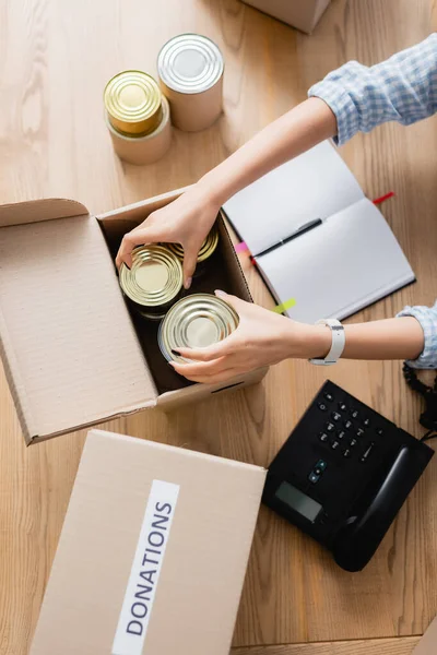 Blick von oben auf freiwillige Helfer, die Konserven in Schachteln neben Notizbuch und Telefon auf dem Tisch verpacken — Stockfoto