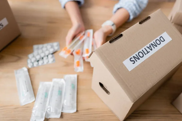 Cropped view of box with donations lettering and volunteer holding syringes near pills on blurred background — Stock Photo