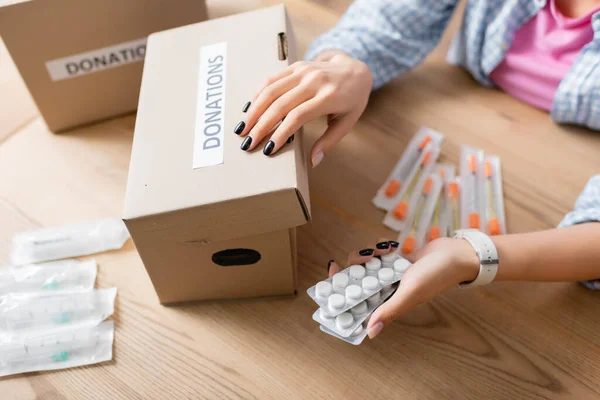 Vista recortada de la mujer sosteniendo píldoras cerca de jeringas y caja con letras de donaciones en la mesa - foto de stock