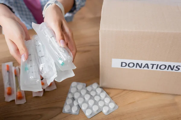 Vista ritagliata di siringhe nelle mani del volontario vicino a pillole e scatola con lettere di donazioni — Foto stock