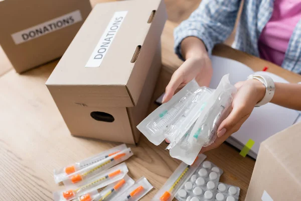 Cropped view of volunteer holding syringes near pills and packages with donations lettering — Stock Photo