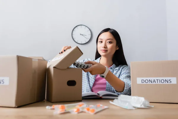 Asian volunteer putting pills in box with donations lettering near notebook and syringes on blurred foreground — Stock Photo