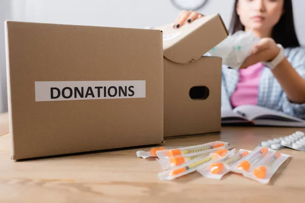 Vista ritagliata della scatola con lettere di donazioni vicino a siringhe, pillole e volontari su sfondo sfocato — Foto stock