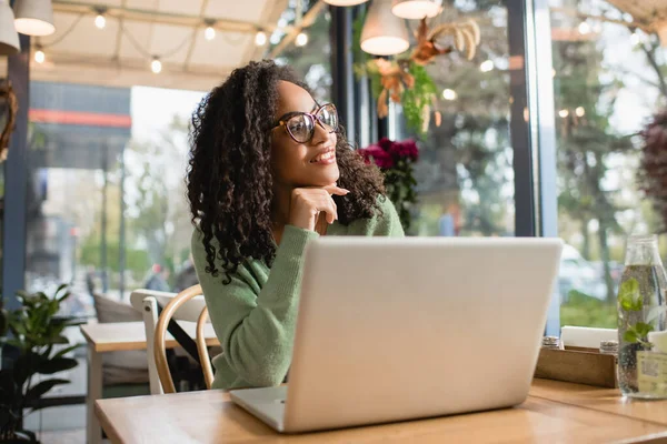 Mujer afroamericana soñadora en gafas sonriendo mientras mira hacia otro lado cerca de la computadora portátil en la mesa - foto de stock