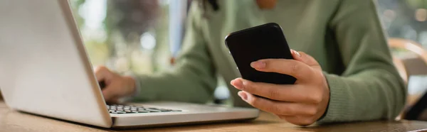 Vista parcial de la mujer afroamericana sosteniendo el teléfono cerca de la computadora portátil en la mesa - foto de stock